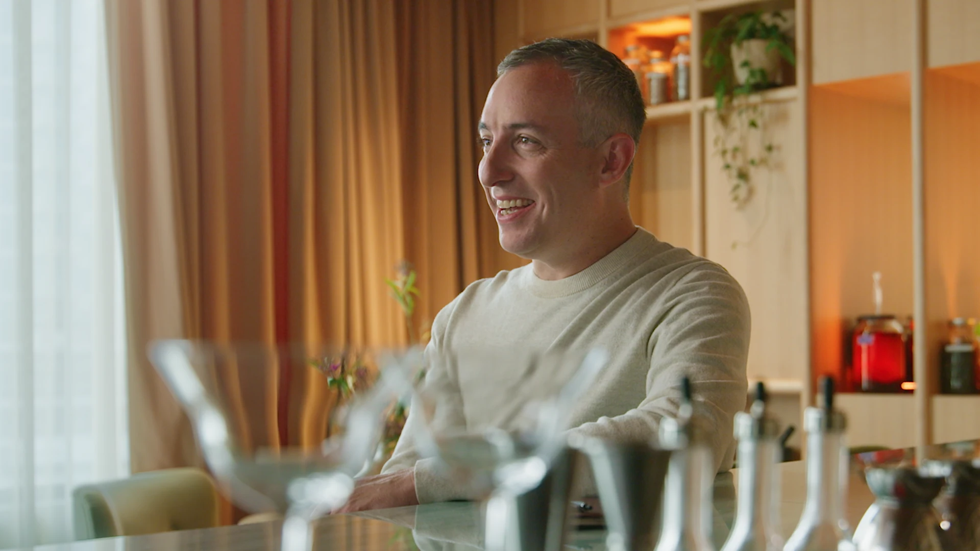 Will Guidara sits at a table, smiling warmly in a softly lit, golden-toned room that feels inviting and full of warmth, with glassware softly blurred in the foreground.